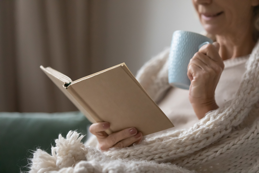 A senior woman reads a book while sipping a hot beverage.